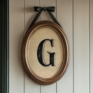 May include: An oval-shaped framed print featuring a large, black letter "G" against a cream-colored background. The frame is gold-toned, and a dark green ribbon is tied at the top for hanging. The artwork is mounted on a white paneled wall.