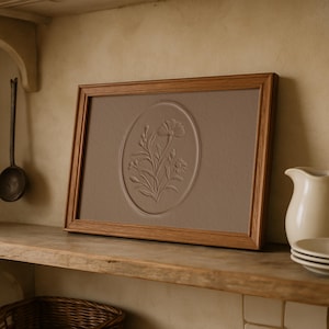 May include: A framed artwork featuring a floral relief in an oval shape. The artwork is set in a light brown frame and rests on a wooden shelf. A white pitcher and plates are visible on the right.