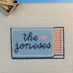 Pode incluir: Uma tela de ponto de agulha com um design de caixa de fósforos azul-claro com o texto "the joneses" em letra azul-escura e um pequeno coração rosa. O design inclui uma fila de cabeças de fósforo laranja e rosa, rodeada por fósforos soltos.