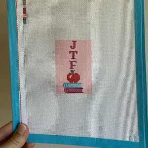 Needlepoint canvas featuring initials and a red apple on a stack of books. This personalized, handmade item is perfect for a bag tag, magnet, paperweight, or framed piece. The initials are fully customizable, making it a thoughtful back-to-school gift, fall or graduation keepsake, or a unique teacher appreciation present.
