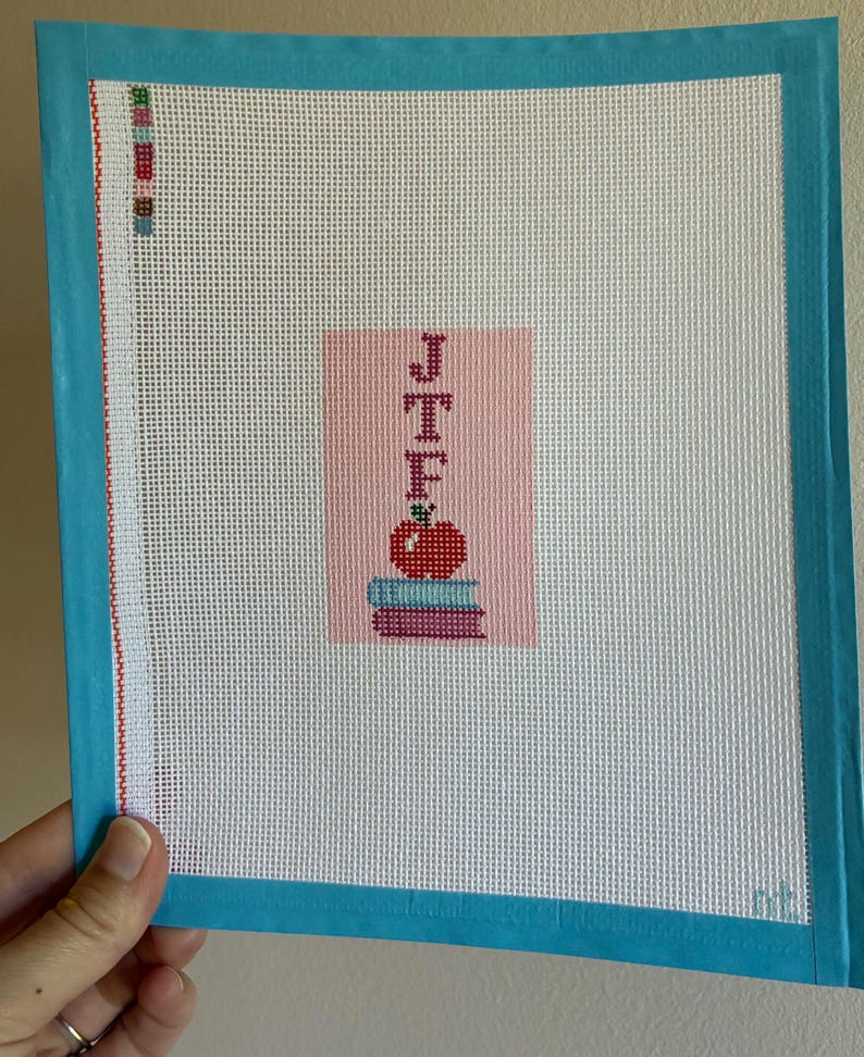 Needlepoint canvas featuring initials and a red apple on a stack of books. This personalized, handmade item is perfect for a bag tag, magnet, paperweight, or framed piece. The initials are fully customizable, making it a thoughtful back-to-school gift, fall or graduation keepsake, or a unique teacher appreciation present.