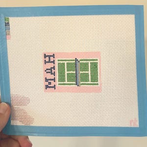 Puede incluir: Lienzo de punto de aguja con un diseño de cancha de tenis y la palabra "HWI" en azul marino. La cancha es verde con una red azul, sobre un fondo rosa. El lienzo tiene un borde azul claro.