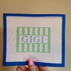 May include: A white canvas with a blue border, featuring a green and white striped design with the word "GIGI" in blue lettering.