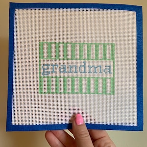 May include: A white canvas with a blue border, featuring a green and blue cross-stitch design that says "grandma".