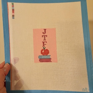 Needlepoint canvas featuring initials and a red apple on a stack of books. This personalized, handmade item is perfect for a bag tag, magnet, paperweight, or framed piece. The initials are fully customizable, making it a thoughtful back-to-school gift, fall or graduation keepsake, or a unique teacher appreciation present.