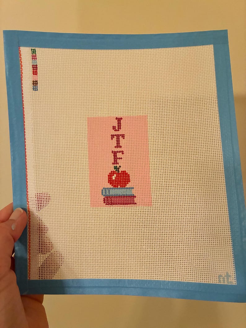 Needlepoint canvas featuring initials and a red apple on a stack of books. This personalized, handmade item is perfect for a bag tag, magnet, paperweight, or framed piece. The initials are fully customizable, making it a thoughtful back-to-school gift, fall or graduation keepsake, or a unique teacher appreciation present.