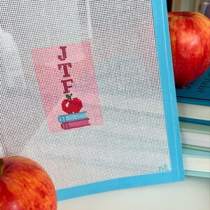 Needlepoint canvas featuring initials and a red apple on a stack of books. This personalized, handmade item is perfect for a bag tag, magnet, paperweight, or framed piece. The initials are fully customizable, making it a thoughtful back-to-school gift, fall or graduation keepsake, or a unique teacher appreciation present.