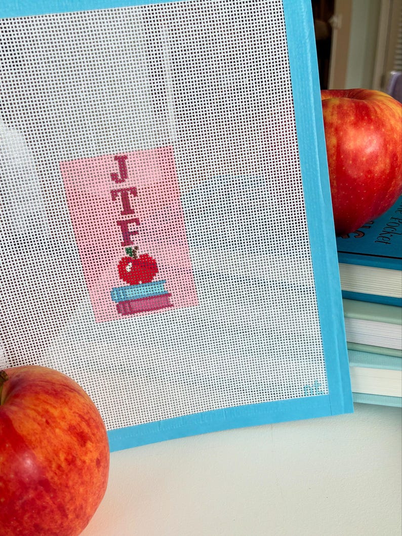 Needlepoint canvas featuring initials and a red apple on a stack of books. This personalized, handmade item is perfect for a bag tag, magnet, paperweight, or framed piece. The initials are fully customizable, making it a thoughtful back-to-school gift, fall or graduation keepsake, or a unique teacher appreciation present.