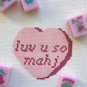Può includere: Un cuore rosa a punto croce con le parole "Iuv u so mahi" in filo rosa scuro. Tessere di Mahjong rosa con simboli e caratteri circondano il cuore. Lo sfondo è un tessuto beige chiaro.