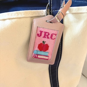 Puede incluir: Una etiqueta de equipaje de cuero rosa con un diseño de punto de cruz con las iniciales "JRC", una manzana roja y una pila de libros. La etiqueta está sujeta a un bolso tote color crema con detalles en azul marino.