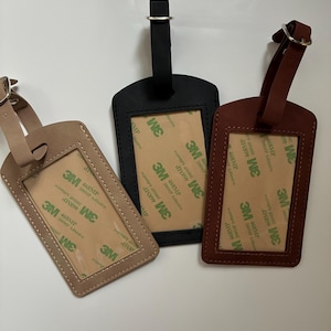 K&ouml;nnte beinhalten: Drei Gep&auml;ckanh&auml;nger aus Leder in Beige, Schwarz und Braun. Jeder Anh&auml;nger hat ein rechteckiges Fenster f&uuml;r eine ID-Karte und einen Riemen mit Metallschnalle. Die Anh&auml;nger sind in verschiedenen Braun- und Schwarzt&ouml;nen gehalten.