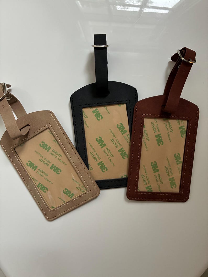 K&ouml;nnte beinhalten: Drei Gep&auml;ckanh&auml;nger aus Leder in Beige, Schwarz und Braun. Jeder Anh&auml;nger hat ein rechteckiges Fenster f&uuml;r eine ID-Karte und einen Riemen mit Metallschnalle. Die Anh&auml;nger sind in verschiedenen Braun- und Schwarzt&ouml;nen gehalten.