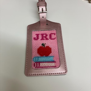 K&ouml;nnte beinhalten: Ein rosa Gep&auml;ckanh&auml;nger mit einem gestickten Design mit den Initialen "JRC", einem roten Apfel und einem Stapel B&uuml;cher. Der Anh&auml;nger hat ein metallisches Finish und einen Schnallenverschluss.