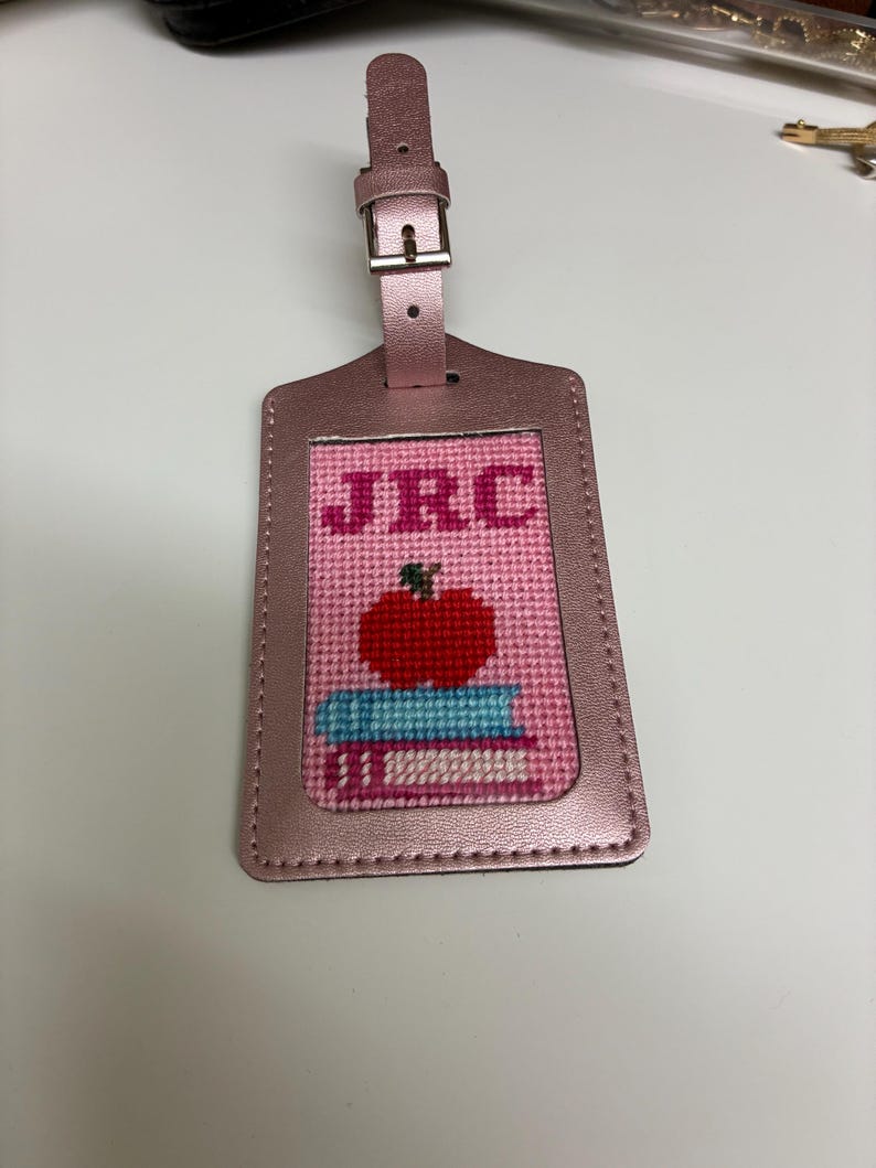 K&ouml;nnte beinhalten: Ein rosa Gep&auml;ckanh&auml;nger mit einem gestickten Design mit den Initialen "JRC", einem roten Apfel und einem Stapel B&uuml;cher. Der Anh&auml;nger hat ein metallisches Finish und einen Schnallenverschluss.