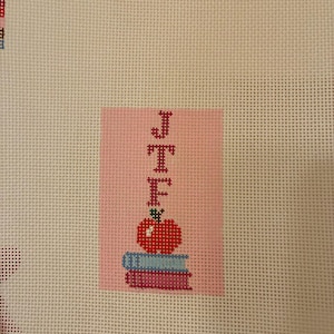 Needlepoint canvas featuring initials and a red apple on a stack of books. This personalized, handmade item is perfect for a bag tag, magnet, paperweight, or framed piece. The initials are fully customizable, making it a thoughtful back-to-school gift, fall or graduation keepsake, or a unique teacher appreciation present.