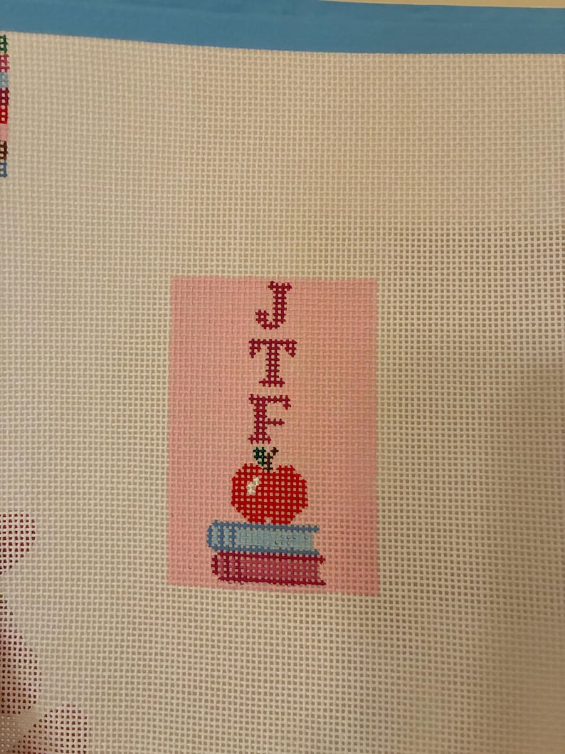 Needlepoint canvas featuring initials and a red apple on a stack of books. This personalized, handmade item is perfect for a bag tag, magnet, paperweight, or framed piece. The initials are fully customizable, making it a thoughtful back-to-school gift, fall or graduation keepsake, or a unique teacher appreciation present.