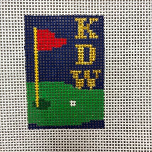 May include: Needlepoint canvas featuring a golf scene. A red flag waves atop a yellow pole on a green putting green. The initials "KDW" are in gold letters against a navy blue background. A white golf hole is visible.