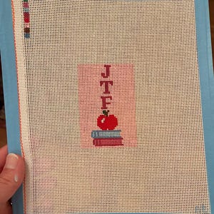 Needlepoint canvas featuring initials and a red apple on a stack of books. This personalized, handmade item is perfect for a bag tag, magnet, paperweight, or framed piece. The initials are fully customizable, making it a thoughtful back-to-school gift, fall or graduation keepsake, or a unique teacher appreciation present.