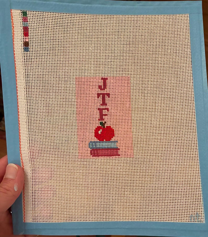 Needlepoint canvas featuring initials and a red apple on a stack of books. This personalized, handmade item is perfect for a bag tag, magnet, paperweight, or framed piece. The initials are fully customizable, making it a thoughtful back-to-school gift, fall or graduation keepsake, or a unique teacher appreciation present.