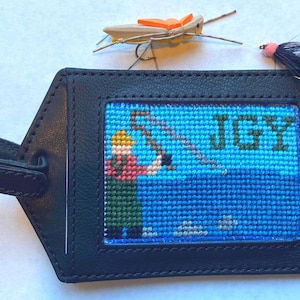 May include: Black leather luggage tag with a stitched design of a person fishing on a blue lake. The tag has the name "JGY" stitched on it.