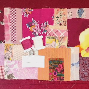 Könnte beinhalten: Ein Patchwork-Textil aus verschiedenen Stoffresten in Rosa-, Rot- und Orangetönen. Das Design umfasst Blumenmuster, Vichy-Karos und Schwanenmotive. Zwei Garnrollen und ein kleines silbernes Objekt sind auf dem Patchwork platziert.