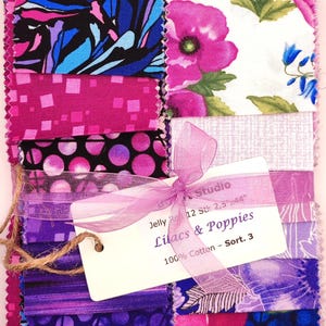 Könnte beinhalten: Ein Stoff-Jelly-Roll-Bundle mit verschiedenen lila, rosa und blauen Blumen- und abstrakten Mustern. Das Bundle ist mit einem lavendelfarbenen Band gebunden und trägt ein Etikett mit der Aufschrift "Lilacs & Poppies, 100% Cotton".