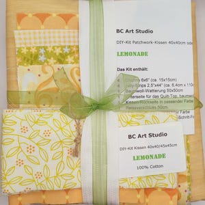 Könnte beinhalten: Ein DIY-Patchwork-Kissen-Set, gebunden mit einem grünen Band. Das Set enthält Stoffstücke in Gelb-, Orange- und Grüntönen mit floralen und geometrischen Mustern. Das Etikett trägt die Aufschrift "LEMONADE" und "100% Cotton".