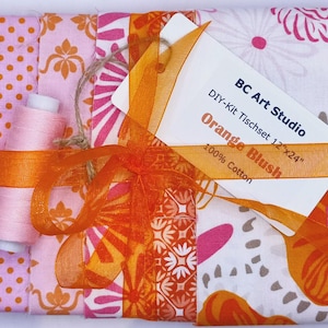 Könnte beinhalten: Ein DIY-Set für ein Tischset mit Stoff in Rosa, Orange und Weiß. Das Set enthält eine Spule rosa Garn und ist mit einem orangefarbenen Band gebunden. Das Etikett lautet "BC Art Studio, Orange Blush, 30cm x 61cm, 100% Baumwolle."