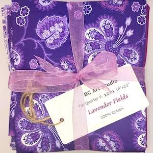 Könnte beinhalten: Ein Bündel aus lila Paisley-Stoffquadraten, gebunden mit einem lavendelfarbenen Band und einem Etikett mit der Aufschrift "Lavender Fields". Der Stoff misst 45.7cm x 55.9cm und besteht aus 100% Baumwolle. Das Design zeigt weiße und lila Blumenmuster.