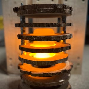 Può includere: Una lampada in legno fatta a mano con un bagliore caldo e invitante. La lampada presenta diverse fette di legno circolari impilate l'una sull'altra, creando un design unico e rustico. La calda luce della lampada brilla attraverso le fette di legno.