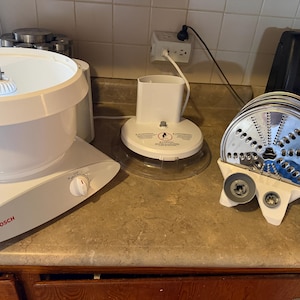 May include: A white Bosch stand mixer with a large mixing bowl, a separate motor base unit with a power cord, and a white plastic stand holding several circular stainless steel slicing and shredding discs with various hole patterns for food prep.