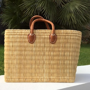 Könnte beinhalten: Eine große, rechteckige geflochtene Strohtasche mit braunen Ledergriffen und Nähten. Die Tasche ist aus natürlichem Stroh gefertigt und hat eine hellbraune Farbe.