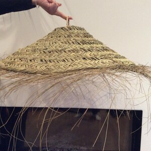 May include: A large, round, woven lampshade made of natural materials. The shade has a fringe of long, thin strands hanging down from the bottom.