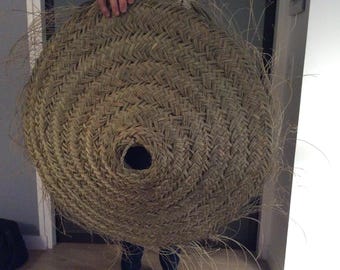 Puede incluir: Una cesta grande, redonda y tejida hecha de materiales naturales. La cesta tiene una abertura circular en el centro y está hecha con un tejido apretado e intrincado.