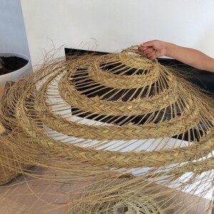 Puede incluir: Un gran sombrero redondo tejido hecho de materiales naturales. El sombrero es marr&oacute;n y tiene un dise&ntilde;o en capas. Est&aacute; siendo sostenido por la mano de una persona.