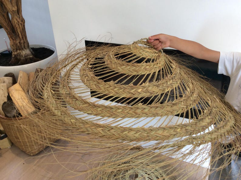 Puede incluir: Un gran sombrero redondo tejido hecho de materiales naturales. El sombrero es marr&oacute;n y tiene un dise&ntilde;o en capas. Est&aacute; siendo sostenido por la mano de una persona.