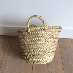 May include: A small, natural woven basket with a single handle. The basket is made of straw and has a light brown colour.