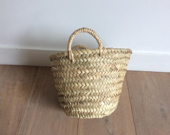 May include: A small, natural woven basket with a single handle. The basket is made of straw and has a light brown color.