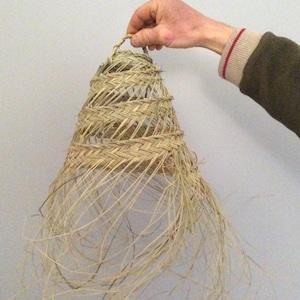 May include: A woven lampshade made of natural straw with a fringed edge. The lampshade is light brown in color and has a conical shape.