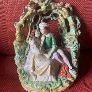Puede incluir: Una figurita de porcelana de una pareja en un abrazo romántico, con un fondo de un árbol estilizado verde y marrón. La mujer lleva un vestido blanco con detalles florales, y el hombre lleva una camisa verde y pantalones marrones.