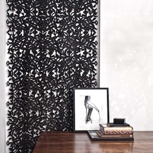 May include: A black metal room divider with a leafy pattern. A wooden desk with a black frame sits in front of the divider. On the desk are a framed black and white drawing, a small black box, and a stack of books with the title "DESTINATIONS" visible.