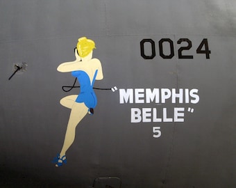 May include: A gray airplane fuselage with a painted image of a woman in a blue dress. The plane is numbered "0024" and has the words "Memphis Belle" and the number "5" painted on it.
