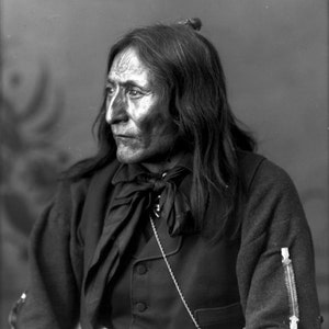 May include: A black and white portrait of a Native American man wearing a dark jacket with a bow tie and a chain with a small silver tag that reads "Standing Bear Chief Ogallala".