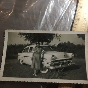 May include: A black and white photograph of a woman standing next to a white 1950s car. The woman is wearing a dress and holding a purse. The car is parked in front of a row of trees.