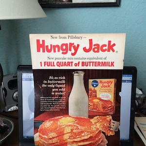 May include: A vintage advertisement for Hungry Jack Buttermilk Pancake Mix. The ad features a stack of pancakes with a bottle of buttermilk and a box of Hungry Jack mix. The text on the ad reads "New from Pillsbury - Hungry Jack. New pancake mix contains equivalent of 1 full quart of buttermilk. It's so rich in buttermilk the only liquid you add is water!"
