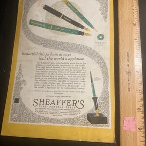 May include: A vintage advertisement for Sheaffer's pens, pencils, and Skrip ink. The ad features a black and green pen with a gold trim and a white dot on the cap. The text reads "Beautiful things have always had the world's applause." and "Sheaffer's Pens, Pencils, Skrip W.A. Sheaffer Pen Company, Fort Madison, Iowa, USA."