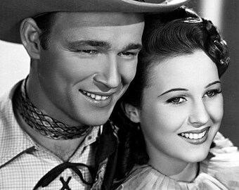 May include: A black and white photo of a man and a woman. The man is wearing a cowboy hat and a plaid shirt. The woman is wearing a white blouse with a ruffled collar. They are both smiling.