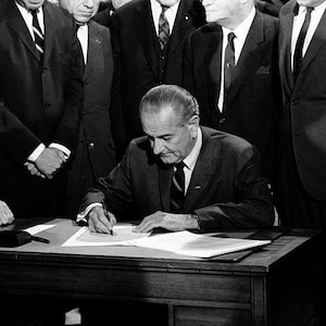 May include: A black and white photo of a man in a suit signing a document at a desk. He is surrounded by other men in suits.