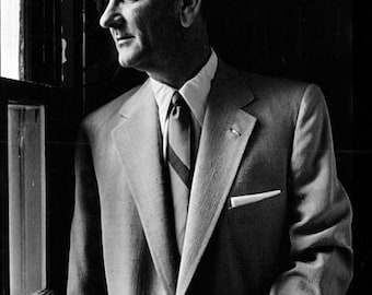May include: Black and white portrait of a man in a light-colored suit, white shirt, and patterned tie, looking out a window. The image is a medium shot, with the man's profile visible. The suit has a pocket square.