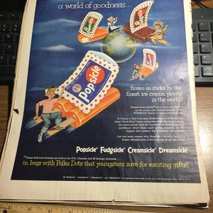 May include: A vintage advertisement for Popsicle, Fudgsicle, Creamsicle, and Dreamsicle frozen treats. The ad features a colorful illustration of the ice cream treats on sticks, with children riding them around a globe. The text reads "a world of goodness... frozen on sticks by the finest ice cream plants in the world!" and "These delicious brands are sold in the U.S., Canada and 40 foreign countries... in bags with Polka Dots that youngsters save for exciting gifts!"